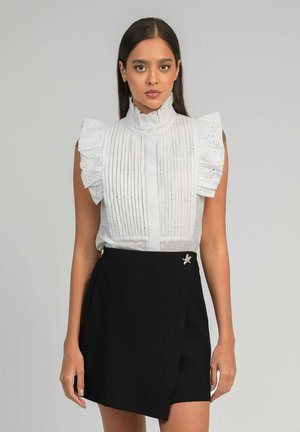 Femme portant un chemisier blanc sans manches à volants avec des plis verticaux et une jupe asymétrique noire ornée d'une épingle décorative en forme d'étoile.