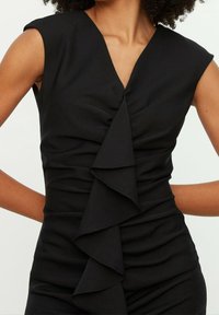 Robe noire avec un décolleté en V profond, des manches courtes et un ruché vertical avec un détail de volants en avant. Tissu lisse et texturé.