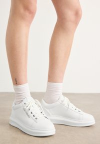 Weiße Leder-Sneaker mit glatter Textur, runder Zehenform und flacher Gummisohle. Die Schnürsenkel sind sichtbar, und die Sockensaum sind weiß.