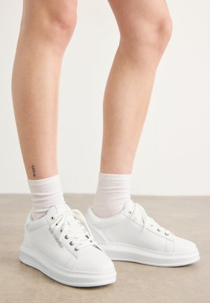 Weiße Leder-Sneaker mit glatter Textur, runder Zehenform und flacher Gummisohle. Die Schnürsenkel sind sichtbar, und die Sockensaum sind weiß.
