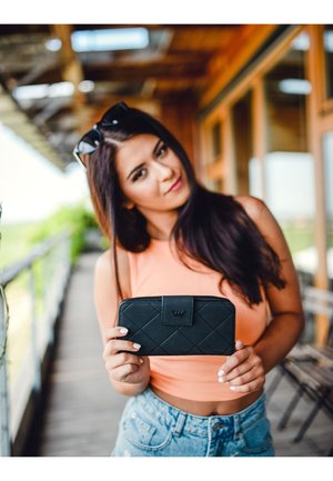 Black quilted wallet with a front flap and logo, held against a light background. The wallet features a textured surface and clean design.