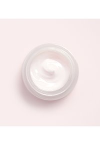 Witte crème in een helder glazen pot met een gladde, gedraaide oppervlak, geplaatst tegen een lichtroze achtergrond.
