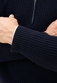 Uomo con le braccia conserte che indossa un maglione a coste di colore blu navy con un polsino bianco visibile e un collo con zip a un quarto.