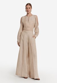 Beige Leinenbluse mit ausgeschnittenen Schultern, Rundhalsausschnitt und Knopfdetail, kombiniert mit weiten beigefarbenen Leinenhosen mit Taschen.