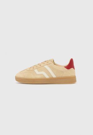 Beige suede sneaker with off-white side stripes, gum sole, red heel tab, and lace-up closure, shown in side profile on white background.
