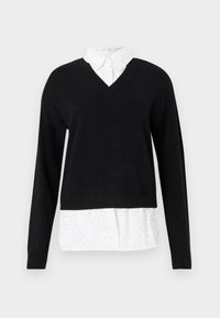 Un pull noir en V superposé sur une chemise blanche à boutons avec un petit motif à pois, présenté sur un fond uni.