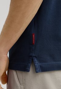 Granatowa koszulka polo z teksturowanym splotem, z rozciętym dołem i czerwoną metką z napisem "JOOP! JEANS" z boku. Widoczne jasne spodnie.