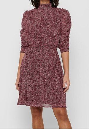 Vrouw draagt een langvallende bordeauxrode bloemenjurk met lange mouwen, een hoge hals, een geplooide taille en pofschouders, staand tegen een effen achtergrond.