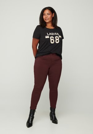 Schwarzes T-Shirt mit weißem "LADIES 68" Druck, kombiniert mit burgunderfarbenen Slim-Fit-Hosen und schwarzen Stiefeletten, stehend vor einem neutralen Hintergrund.