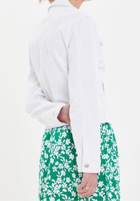 Chaqueta de mezclilla blanca con cuello, mangas largas y herrajes plateados, llevada sobre una falda verde con estampado floral. Tejido texturizado, ajuste casual.