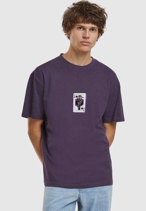 Ung mand iført en mørkelilla T-shirt med et joker-spillekort design og lyseblå jeans, stående med hænderne bag ryggen.