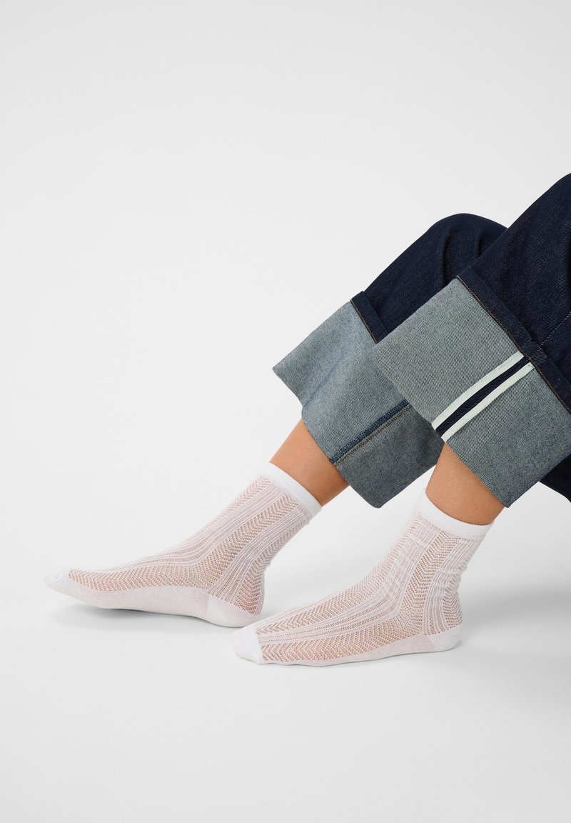 Pieds portant des chaussettes en résille blanches à motifs avec un jean en denim bleu foncé retroussé, sur un fond clair uni.