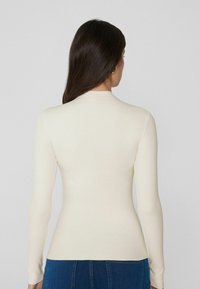 Langsleeve, getailleerde top in crème, met een hoge halslijn en een gladde textuur, gecombineerd met blauwe high-waisted jeans.