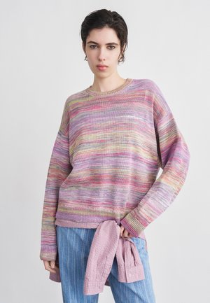 Persona joven con jersey de rayas multicolores, vaqueros azules y una camisa rosa texturizada atada a la cintura, de pie frente a un fondo liso.