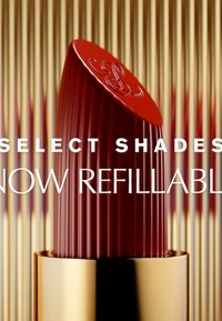 Roter Lippenstift mit gerilltem Design, goldener Hülle und glänzender Oberfläche. Der Text lautet "Ausgewählte Farbtöne jetzt nachfüllbar." Der Hintergrund zeigt vertikale Linien.