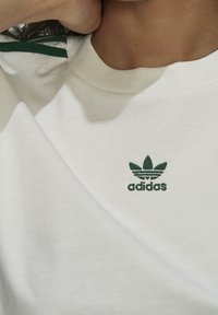 Vit bomulls t-shirt med grön broderad Adidas-logotyp på bröstet och gröna ränder på övre axlar. Mjuk, avslappnad design.