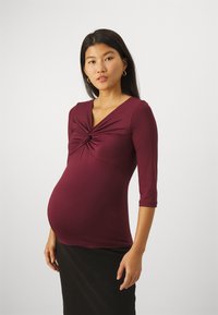 Maternity top in burgundy, made of stretchy fabric. Features a twisted design at the front and 3/4 length sleeves. Paired with a black skirt.