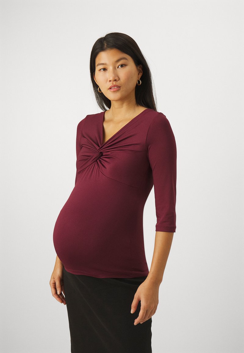 Maternity top in burgundy, made of stretchy fabric. Features a twisted design at the front and 3/4 length sleeves. Paired with a black skirt.