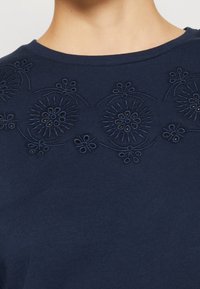 Top in cotone blu navy con ricami floreali al colletto, con cuciture a contrasto e forma classica del collo a giro.