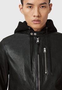 Veste en cuir noir avec une texture lisse, dotée d'un col haut, de zips argentés et d'une capuche. Fermeture éclair à l'avant et détails réglables.