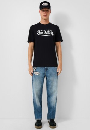 Mężczyzna stoi na szarym tle, ubrany w czapkę Von Dutch w kolorze czarnym, czarną koszulkę Von Dutch, niebieskie dżinsy z łatami i czarne sneakersy.