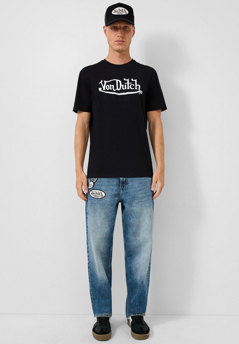 Muž stojí proti šedému pozadí, nosí černou čepici Von Dutch, černé tričko Von Dutch, modré džíny s nášivkami a černé tenisky.