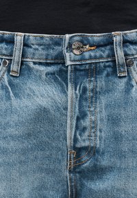 Κοντινή λήψη στημένος της μέσης ενός μπλε τζιν με μεταλλικό κουμπί που γράφει "DENIM" και εμφανείς ραφές στην τσέπη και τις θηλιές της ζώνης.