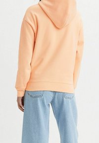 Felpa con cappuccio color pesca dalla texture morbida, con vestibilità ampia e polsini a costine, abbinata a jeans a gamba larga di colore azzurro chiaro.