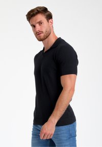 Melna polo krekls ar ribota tekstūra, īsām piedurknēm un klasisku apkakli, apvienots ar gaiši zilu džinsu biksēm.