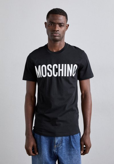 Crna pamučna majica s podebljanim bijelim natpisom "MOSCHINO" sprijeda, u kombinaciji s širokim plavim trapericama.