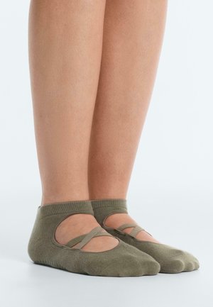 Pár nôh v olivovo zelených baletných ponožkách s krížovými elastickými remienkami, stojacich na svetlosivej ploche.