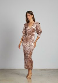 Donna che indossa un lungo abito aderente in velluto mauve con maniche a sbuffo a tre quarti e una profonda scollatura a V, in piedi su un pavimento in cemento semplice.