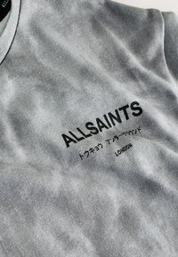 Šedá látka s čiernym textom „ALLSAINTS“, japonskými znakmi a nápisom „LONDON“ na zloženej odevnej látke.