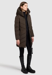 Cappotto imbottito marrone con colletto alto e cappuccio, caratterizzato da un design a cuciture diagonali. Indossato su leggings neri e stivali neri fino al ginocchio.