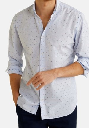 Homme portant une chemise bleu clair à boutons avec de petits points colorés, manches retroussées, une main dans la poche et l'autre pointant vers l'avant.