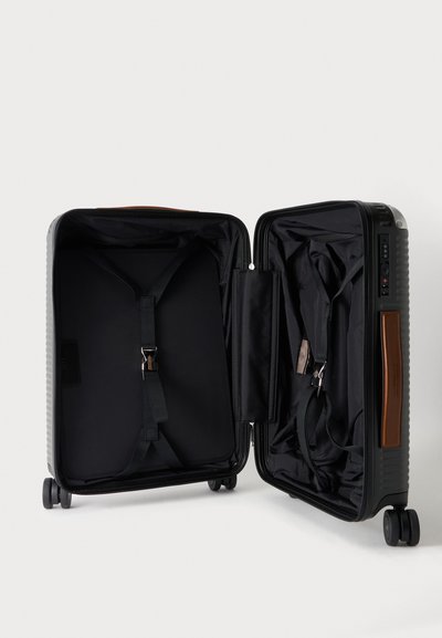 Valise rigide noire ouverte avec des sangles intérieures, des compartiments zippés, quatre roues et une poignée marron sur fond blanc.