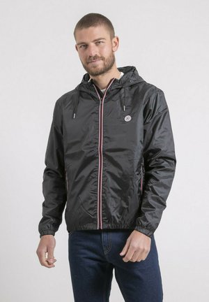 Homme portant une veste à capuche noire zippée et un jean bleu, debout devant un fond clair uni, avec un léger sourire.