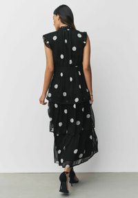 Schwarzes, gestuftes Kleid mit weißen Pünktchen, transparentem Stoff, Rüschensäumen und tailliertem Schnitt. Getragen mit schwarzen High Heels.