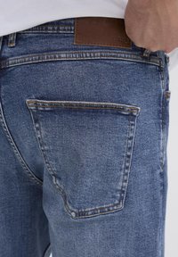 Jean en denim bleu avec une poche arrière, étiquette en cuir brun cousue à la taille, avec des coutures contrastées et une texture décolorée.