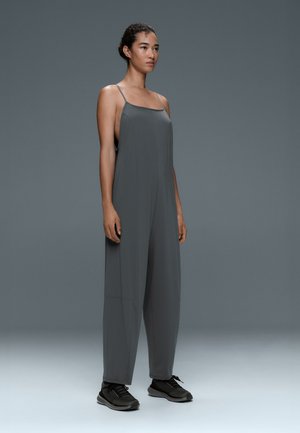Femme debout portant une combinaison ample sans manches gris foncé avec de fines bretelles et des baskets noires sur un fond gris uni.