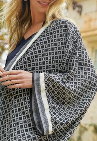Zwarte kimono-achtige wikkel met een wit geometrisch patroon, contrasterende witte en gemusterde afwerking. Zachte stof met losse, vloeiende mouwen.