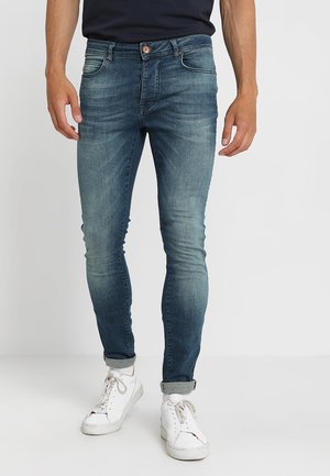 Strakke blauwe spijkerbroek met een vervaagde afwerking, vijf zakken en een koperen knoopsluiting. Draagt zich met witte sneakers.