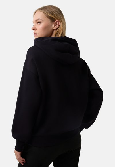 Frau mit blonden Haaren, die einen lockeren schwarzen Kapuzenpullover trägt, von hinten gezeigt und zur Seite blickend vor einem weißen Hintergrund.