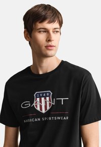 Młody mężczyzna w czarnej koszulce GANT z czerwono-białą pasiastą tarczą i napisem „1949 American Sportswear”, patrzący na bok na szarym tle.
