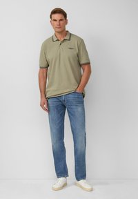 Oliwkowa koszulka polo z czarno-białym kołnierzykiem, zestawiona z jasnoniebieskimi dżinsami i białymi sneakersami. Prosty, casualowy styl.