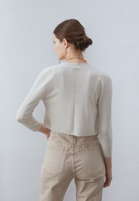 Világosszürke, cropped top háromnegyedes ujjakkal; textúrált kötött anyag, amely enyhe fényt tükröz; hátsó varrás részlet; bézs, magas derekú nadrággal párosítva.
