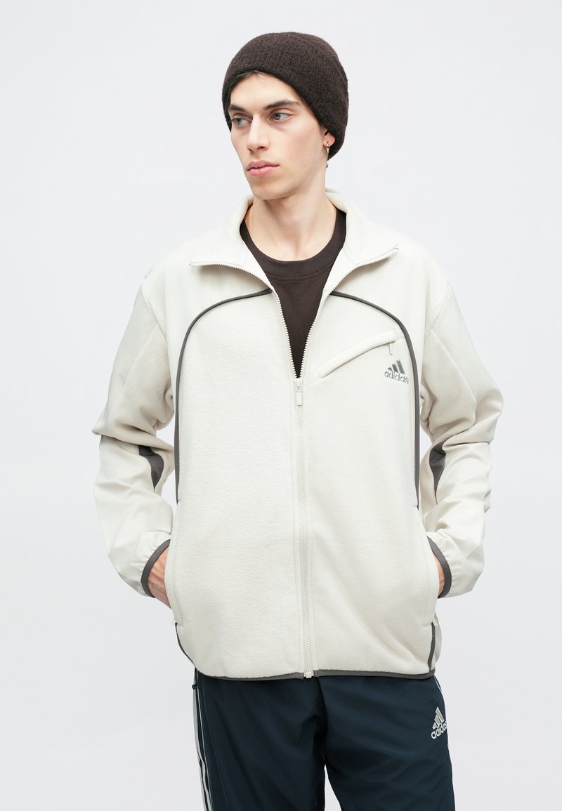 Licht beige fleece jacket met zwarte accenten, ritssluiting aan de voorkant en logo op de borst. Het model draagt een donkere beanie en staat met de handen in de zakken.