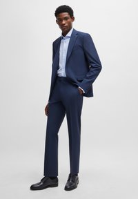 Marineblauw pak met gestructureerde schouders en slanke pasvorm. Bevat een lichtblauw overhemd en zwarte veterschoenen. Gladde stoftextuur.