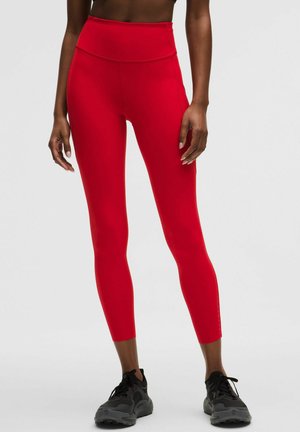 Naine, kes kannab erksaid punaseid kõrge vöökohaga lühikesi retuuse ja musti spordijalatseid, seisab ühevärvilise heleda tausta ees.