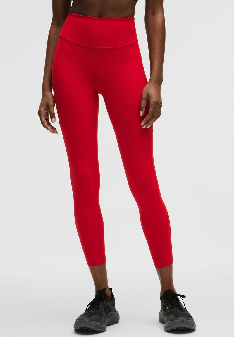 Kvinde iført skrigrøde, højtaljede, korte leggings og sorte atletiksko, stående mod en ensfarvet lys baggrund.